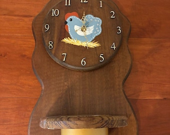 Op de afbeelding: Een houten wandklok met een donkerbruine afwerking en een geschulpte rand. De wijzerplaat heeft goudkleurige cijfers, gouden wijzers en een geschilderde blauwe kip in een geel nest. Een goudkleurige metalen beker is eronder bevestigd.