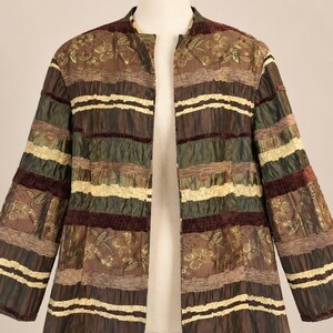 May include: A long-sleeved jacket featuring a horizontal striped pattern in shades of brown, olive green, and cream. The garment includes textured fabric panels, decorative trim, and a subtle floral print on the brown sections, with an open front.