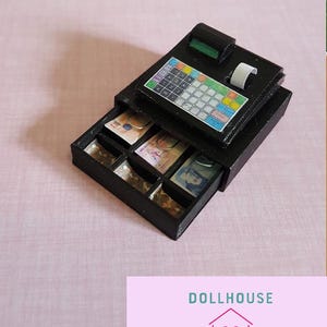 May include: A black cash register with a drawer that opens to reveal compartments for coins and bills. The cash register has a digital display and a keypad with colourful buttons.