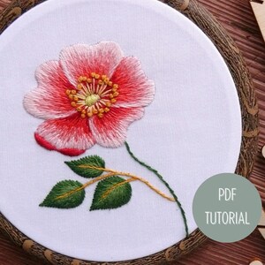 以下が含まれることがあります： 白い布に刺繍された花の模様が、丸い刺繍枠に入っています。花はピンクと赤で、緑の葉が付いています。枠は木製の表面に置かれ、様々な色の刺繍糸が置かれています。
