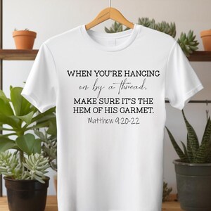 May include: White t-shirt with black text. The text reads "WHEN YOU'RE HANGING on by a thread. MAKE SURE IT'S THE HEM OF HIS GARMENT. Matthew 9:20-22". The shirt is hanging on a wooden hanger.