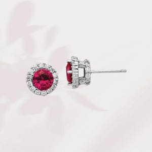 May include: A pair of silver stud earrings with a red gemstone surrounded by a halo of smaller white gemstones.