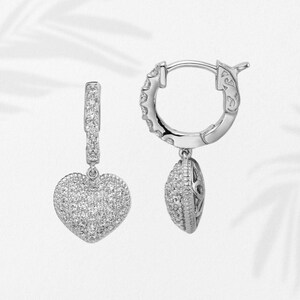May include: A pair of silver hoop earrings with heart-shaped pendants. The earrings are encrusted with small, sparkling stones. The heart pendants are fully covered in the stones, and the hoops have a decorative design.