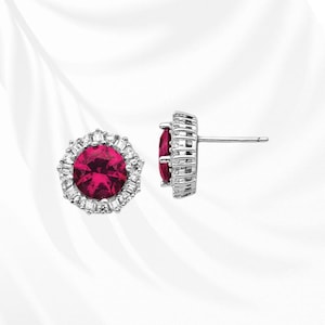 May include: A pair of stud earrings featuring a large, round, deep red gemstone surrounded by a halo of smaller, clear gemstones. The earrings have a silver-colored metal setting and a post back.