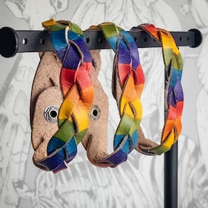 Op de afbeelding: Gevlochten leren armbanden in regenboogkleuren, tentoongesteld op een zwart metalen rek. De armbanden zijn gemaakt van geweven leren stroken, met rode, oranje, gele, groene, blauwe en paarse tinten. De achtergrond heeft een neutraal patroon.