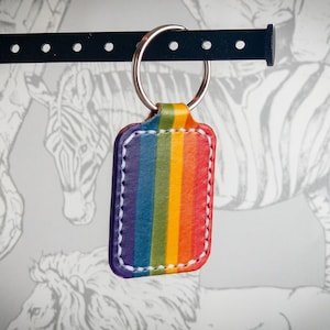 Op de afbeelding: Een rechthoekige leren sleutelhanger met een regenboogstreepontwerp. De sleutelhanger heeft een zilveren metalen ring en witte stiksels langs de randen. De achtergrond heeft een grijs-witte dierenprint.