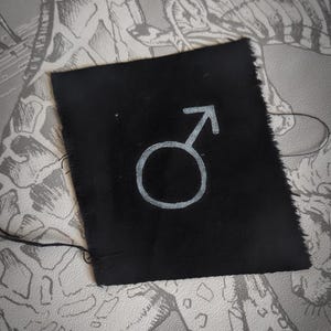 Op de afbeelding: Een klein, vierkant zwart stoffen lapje met een zilverkleurig symbool van Mars, een cirkel met een pijl die naar boven en rechts wijst. De randen van het lapje zijn onafgewerkt. De achtergrond is een patroon, grijs en wit oppervlak.