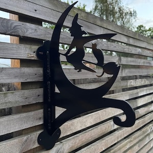 May include: Black metal silhouette of a witch on a broom with a cat, set against a crescent moon design. The artwork is mounted on a wooden fence. A decorative Halloween or autumn garden ornament.