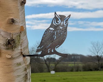 May include: A dark grey metal owl silhouette featuring cut-out details for the eyes, feathers, and wings. The owl is perched on a horizontal branch extension that is inserted directly into the side of a light-coloured birch tree trunk.