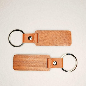 Pode incluir: Dois chaveiros retangulares de madeira com uma tira de couro castanho claro e um porta-chaves prateado. A madeira tem um padrão de grão natural. A tira de couro é presa com um botão de pressão prateado.