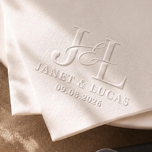May include: A white paper napkin featuring an embossed monogram with the letters J and L, followed by the text JANET & LUCAS and the date 09.08.2026. The napkin is folded and rests on a white plate, with a metal embossing tool visible nearby.