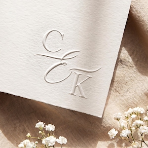 May include: A thick, off-white textured paper card featuring a blind embossed monogram of the letters C and K in a cursive script. The card is positioned against a beige linen fabric background with small white baby's breath flowers nearby.