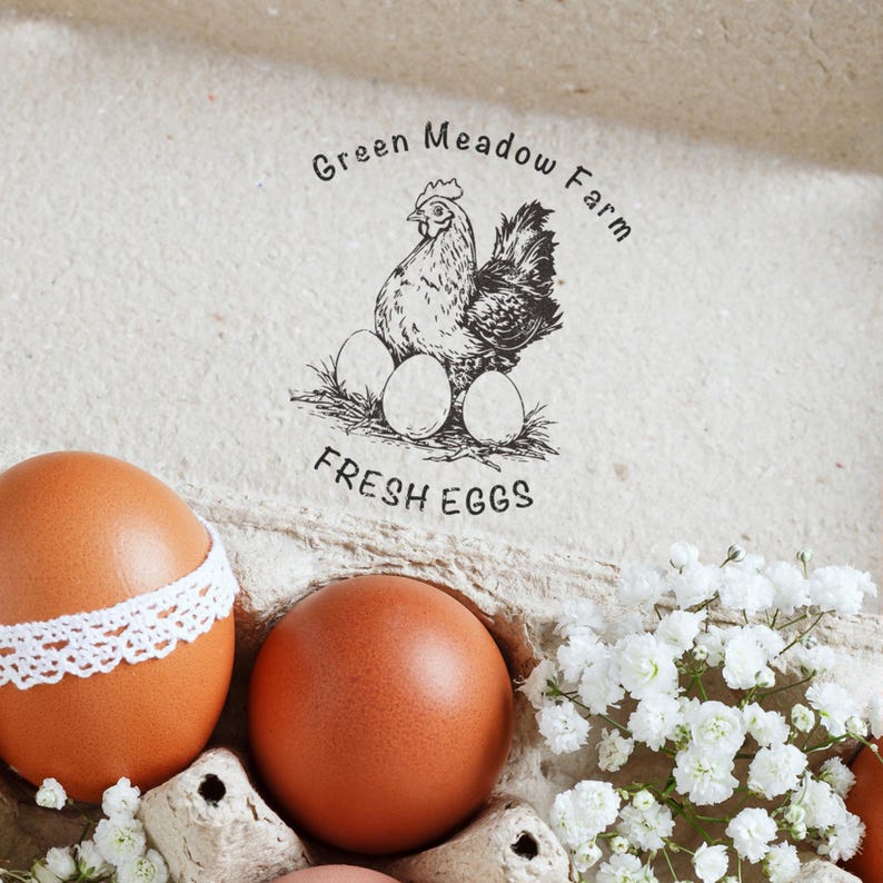 May include: A close-up of brown eggs, some with white lace trim, in a carton with white flowers. The image features a stamp with a hen, eggs, and the text "Green Meadow Farm FRESH EGGS."