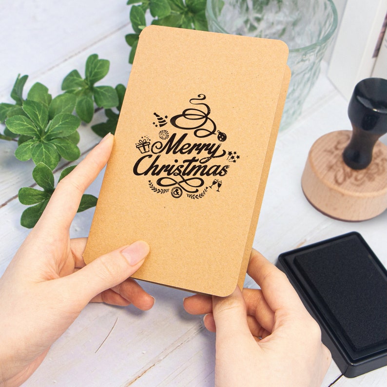 May include: A brown card with a black rubber stamp that says "Merry Christmas" with a festive design. The stamp is being held by a person's hands.