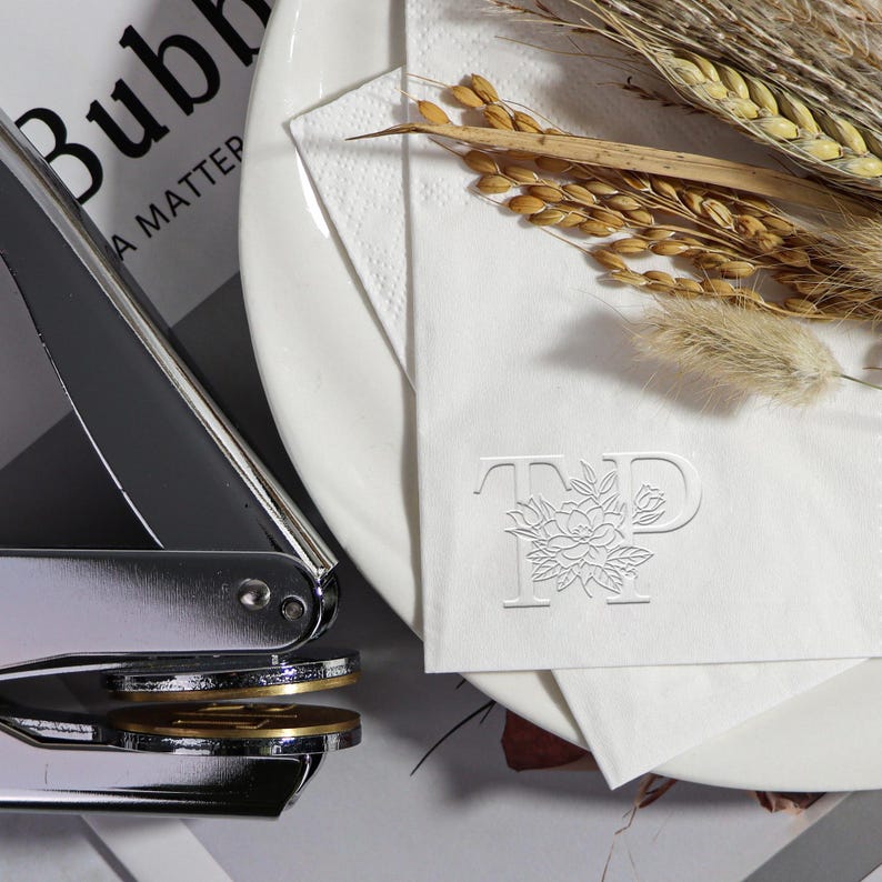 May include: A close-up of a white napkin with a custom embossed design of the letters "TP" with a floral motif. The napkin is on a white plate with dried wheat stalks and a silver stamp.