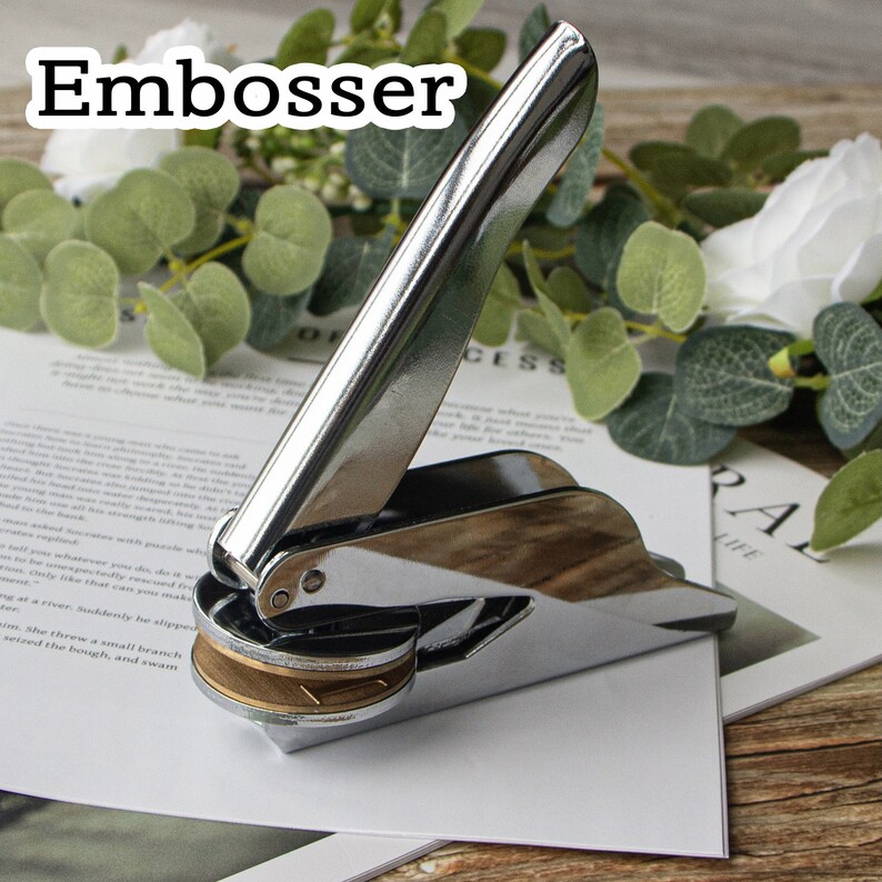May include: A silver metal embosser with a handle and a round, gold-colored embossing plate. The embosser is on a white piece of paper, which is on a wooden surface.