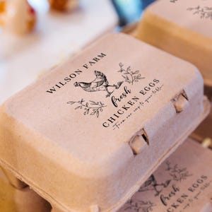 May include: A close-up of a cardboard egg carton with the text "WILSON FARM" and "fresh CHICKEN EGGS" printed in black. The carton features a drawing of a chicken and floral accents. The phrase "From our coop to your table" is also printed on the carton.