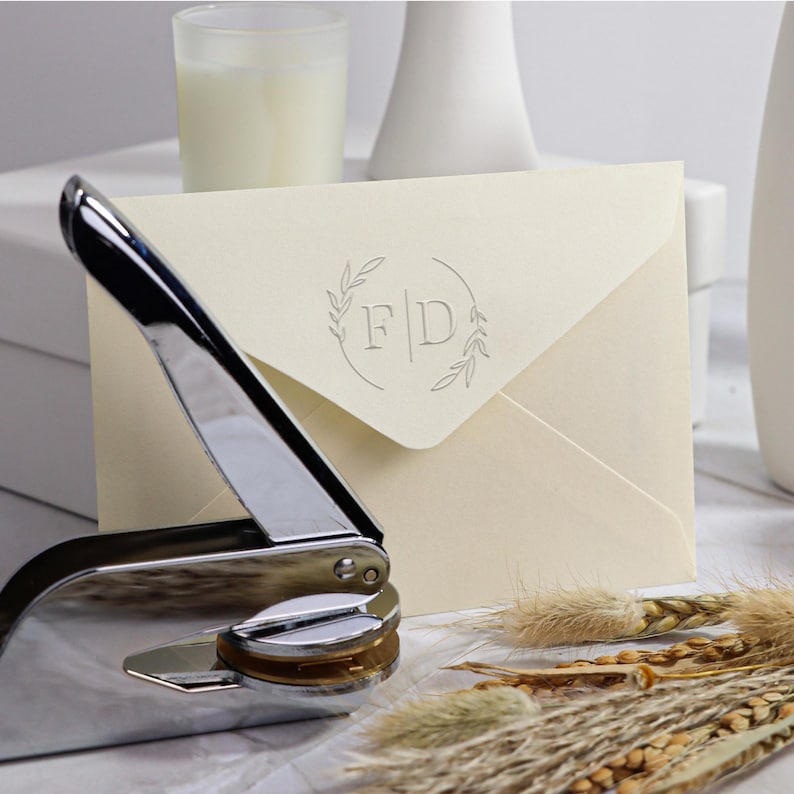 May include: A silver self-inking stamp with a round design featuring the letters "F|D" surrounded by a wreath of leaves. The stamp is on a cream envelope.