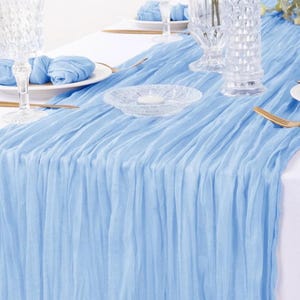 Op de afbeelding: Een lichtblauwe geplooide tafelrunner. De runner is gemaakt van een zachte, vloeiende stof en heeft een subtiele glans. Het is perfect om een ​​toets van elegantie toe te voegen aan elke tafelsetting.