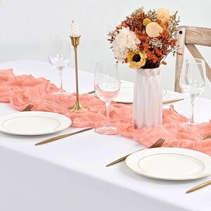 May include: A coral pink table runner with a ruffled edge is laid on a white tablecloth. A gold candlestick, two wine glasses, and two white plates are set on the table. A white vase with a bouquet of dried flowers is in the center of the table.