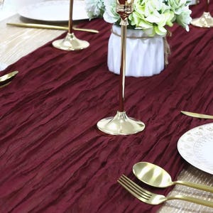 Puede incluir: Un camino de mesa de terciopelo aplastado burdeos con candelabros dorados y un jarrón blanco con flores verdes.