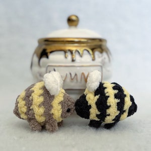 May include: Two crocheted bee plushies, one with yellow and brown stripes and the other with black and yellow stripes, are facing each other. The plushies have white wings and are in front of a white ceramic honey pot with gold accents.