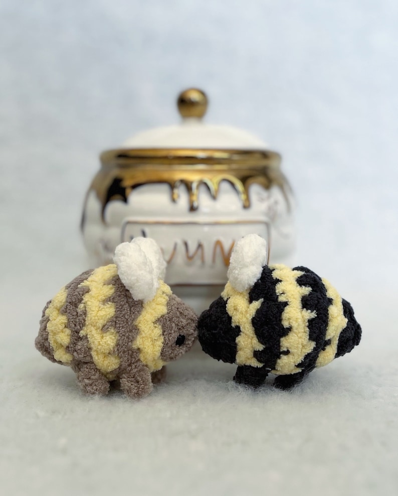 May include: Two crocheted bee plushies, one with yellow and brown stripes and the other with black and yellow stripes, are facing each other. The plushies have white wings and are in front of a white ceramic honey pot with gold accents.