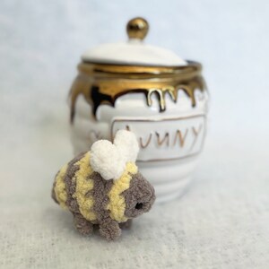 May include: A small, crocheted bee toy with yellow and brown stripes and white wings. The bee is sitting in front of a white ceramic honey pot with gold accents and the word "Honey" written on it.