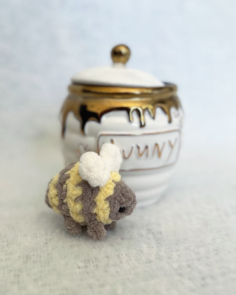 May include: A small, crocheted bee toy with yellow and brown stripes and white wings. The bee is sitting in front of a white ceramic honey pot with gold accents and the word "Honey" written on it.