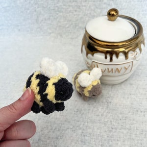 May include: Two crocheted bumblebees, one yellow and black, the other brown and yellow, with white wings. The bees are sitting in front of a white ceramic honey pot with gold accents.