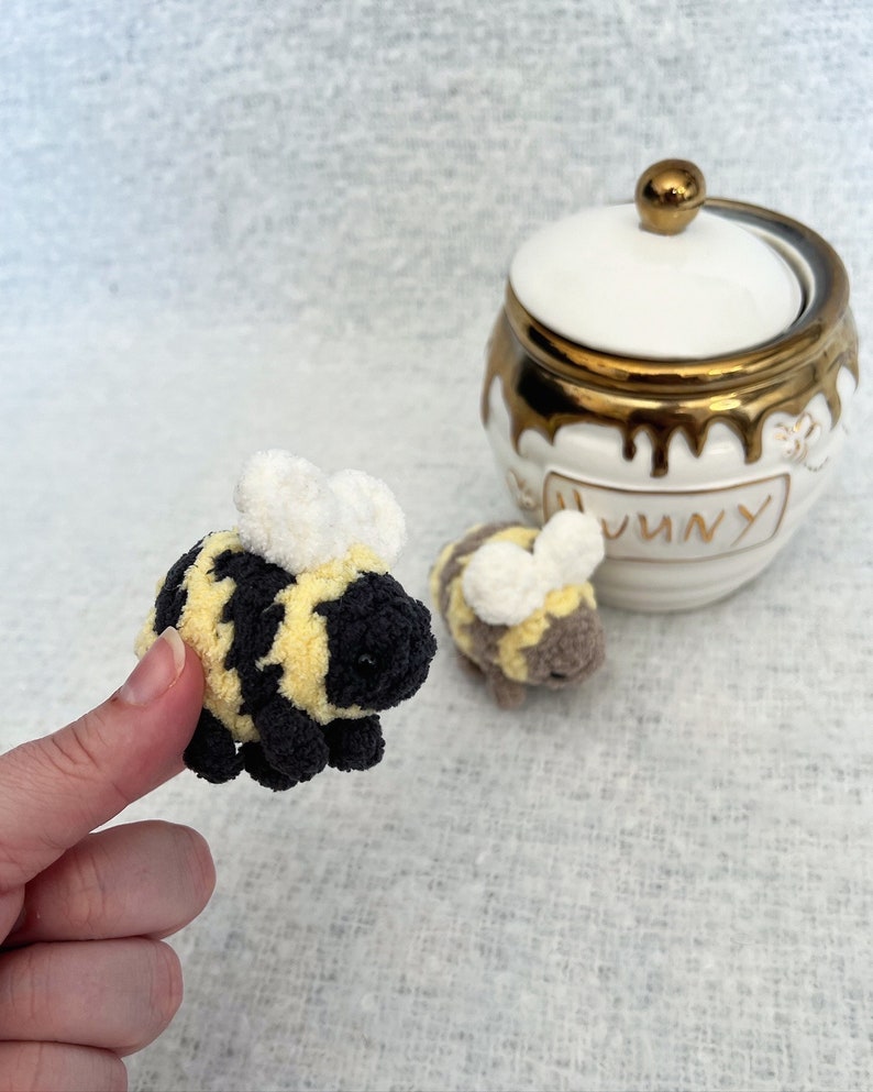 May include: Two crocheted bumblebees, one yellow and black, the other brown and yellow, with white wings. The bees are sitting in front of a white ceramic honey pot with gold accents.
