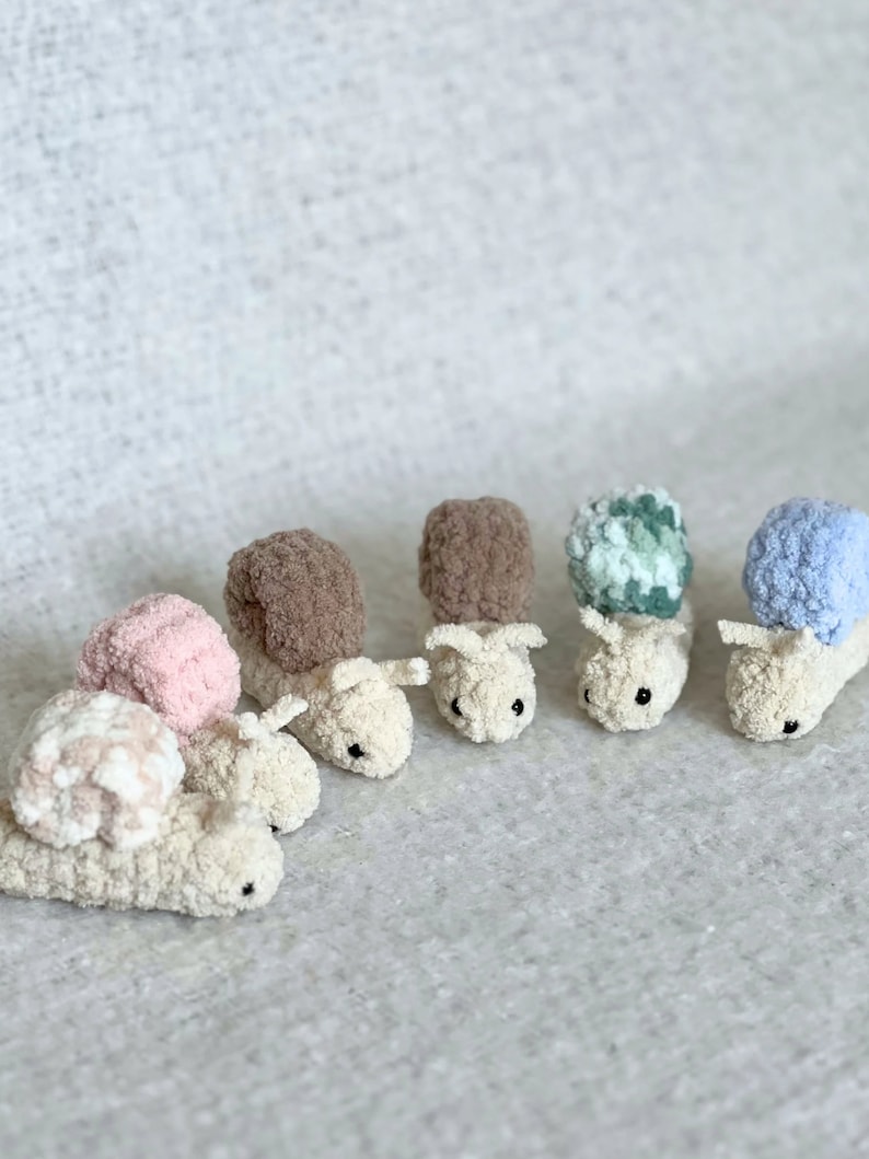 Puede incluir: Cinco juguetes de caracol de crochet en diferentes colores, incluyendo rosa, marr&oacute;n, verde, azul y blanco. Los caracoles est&aacute;n todos mirando en la misma direcci&oacute;n y tienen ojos negros.