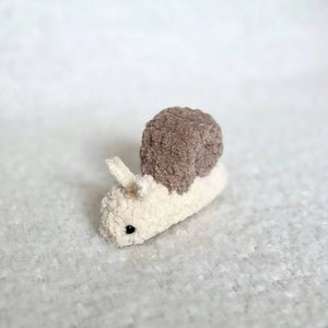 Puede incluir: Un peque&ntilde;o juguete de peluche en forma de caracol con una concha marr&oacute;n y un cuerpo blanco. El caracol tiene un ojo negro y est&aacute; hecho de tela suave y peluda.