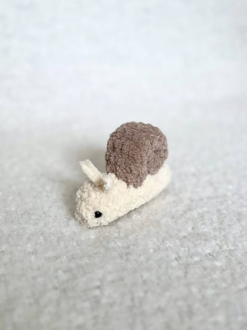 Puede incluir: Un peque&ntilde;o juguete de peluche en forma de caracol con una concha marr&oacute;n y un cuerpo blanco. El caracol tiene un ojo negro y est&aacute; hecho de tela suave y peluda.