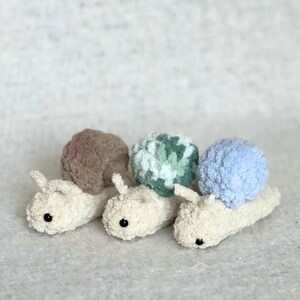 Puede incluir: Tres juguetes de caracol de crochet con conchas de diferentes colores. Los caracoles son blancos con ojos negros y est&aacute;n alineados en una fila. Las conchas son marrones, verdes y blancas, y azul claro.