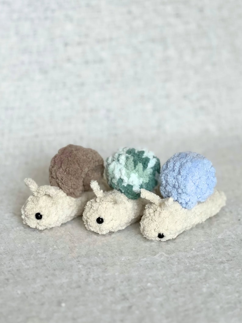 Puede incluir: Tres juguetes de caracol de crochet con conchas de diferentes colores. Los caracoles son blancos con ojos negros y est&aacute;n alineados en una fila. Las conchas son marrones, verdes y blancas, y azul claro.