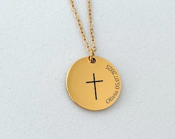May include: Gold-colored necklace with a round pendant featuring a black cross and engraved text. The chain is delicate, and the pendant is centered. The necklace rests on a white surface.