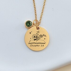 Op de afbeelding: Goudkleurige ketting met een ronde hanger met een bloemenontwerp en de tekst "Chapter 18". Een kleine groene edelsteen hangt in de buurt van de hanger. De ketting wordt gepresenteerd op een wit oppervlak.