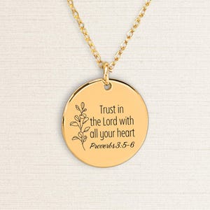 Könnte beinhalten: Goldfarbene Halskette mit Anhänger und rundem Charm. Der Charm trägt den Text "Trust in the Lord with all your heart Proverbs 3:5-6" und ein florales Design. Die Halskette wird vor hellem Hintergrund gezeigt.