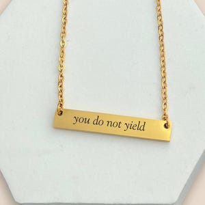 May include: Gold-colored bar necklace with the inscription "you do not yield" in a simple, sans-serif font. The necklace hangs on a delicate chain. The background is a white, geometric shape.