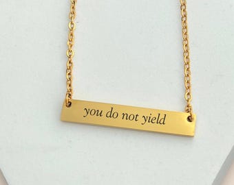 May include: Gold-colored bar necklace with the inscription "you do not yield" in a simple, sans-serif font. The necklace hangs on a delicate chain. The background is a white, geometric shape.