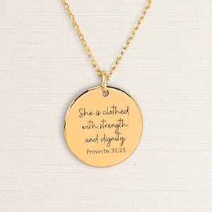 Könnte beinhalten: Eine goldfarbene Anhängerkette. Der runde Anhänger trägt die Inschrift "She is clothed with strength and dignity" und "Proverbs 31:25" in schwarzer Schrift. Die Halskette hat eine zarte Kette.