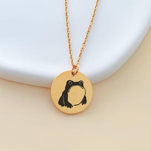 Puede incluir: Un collar de tono dorado con un colgante circular que presenta una ilustración grabada en negro de una rana. El colgante cuelga de una delicada cadena de tono dorado, mostrando un diseño sencillo con un gráfico claro del anfibio.