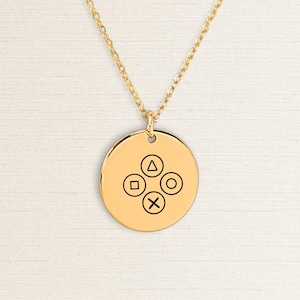 May include: A gold-colored pendant necklace featuring a circular charm with four black game controller symbols: a triangle, square, circle, and X. The pendant hangs from a delicate gold chain, set against a neutral background.