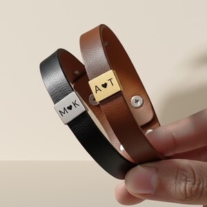 Pode incluir: Duas pulseiras de couro, uma preta e outra castanha, com detalhes metálicos personalizados. A pulseira preta tem gravado "M ♥ K", enquanto a castanha exibe "A ♥ T". As pulseiras são presas com parafusos prateados.