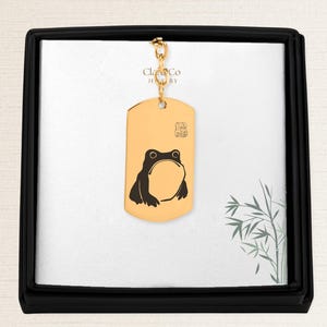 May include: Gold-colored dog tag pendant with a black frog illustration. The pendant hangs from a gold chain and is presented in a black box with a white interior. The brand name "Clara Co Jewelry" is visible.