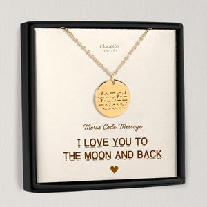 May include: Gold-coloured necklace with a round pendant featuring a Morse code message. The necklace is displayed in a black box with the text "I LOVE YOU TO THE MOON AND BACK" in brown. The brand name "ClaralCo JEWELRY" is visible.