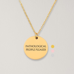 May include: A gold-toned necklace featuring a circular pendant with the black engraved text PATHOLOGICAL PEOPLE PLEASER. The pendant hangs from a delicate gold-toned chain. To the right of the pendant are three small colored circles in gold, pink, and silver.