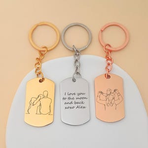 Op de afbeelding: Drie metalen sleutelhangers met dog tag-hangers. Eén is goudkleurig met een illustratie van een stel, één is zilverkleurig met tekst en één is roségoudkleurig met een familie-illustratie. Elk heeft een ring en een ketting.