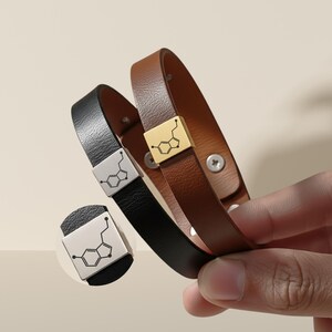 May include: Two leather bracelets, one black and one brown, each with a metal clasp displaying a serotonin molecule design. The brown bracelet has a gold clasp, the black one a silver clasp. They fasten with a snap closure.